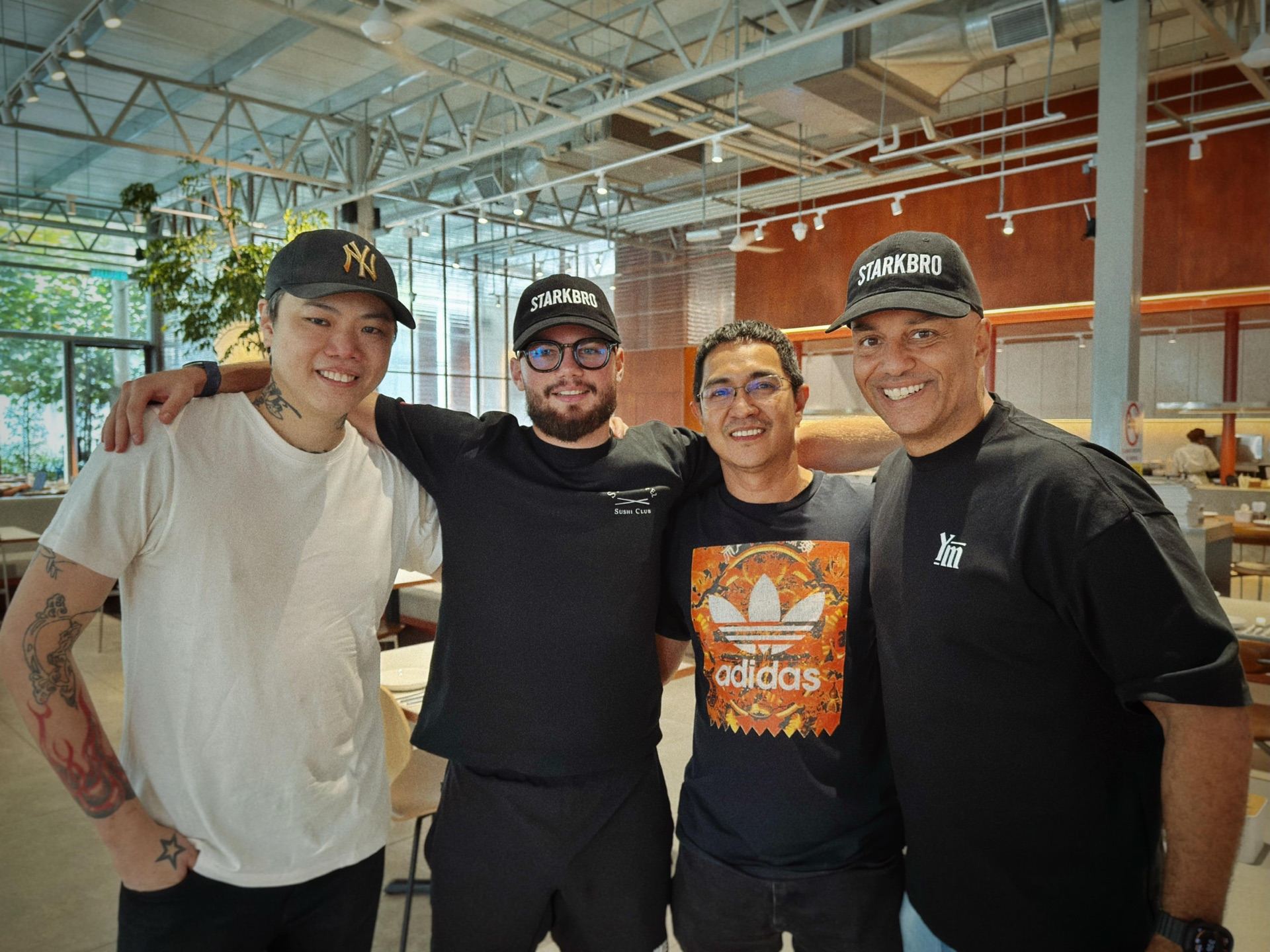 Four friends arm in arm in a bright restaurant, two wearing STARKBRO caps