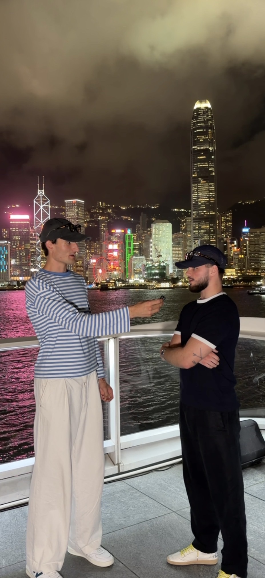 Two friends in caps talking in front of the Hongkong skyline at night